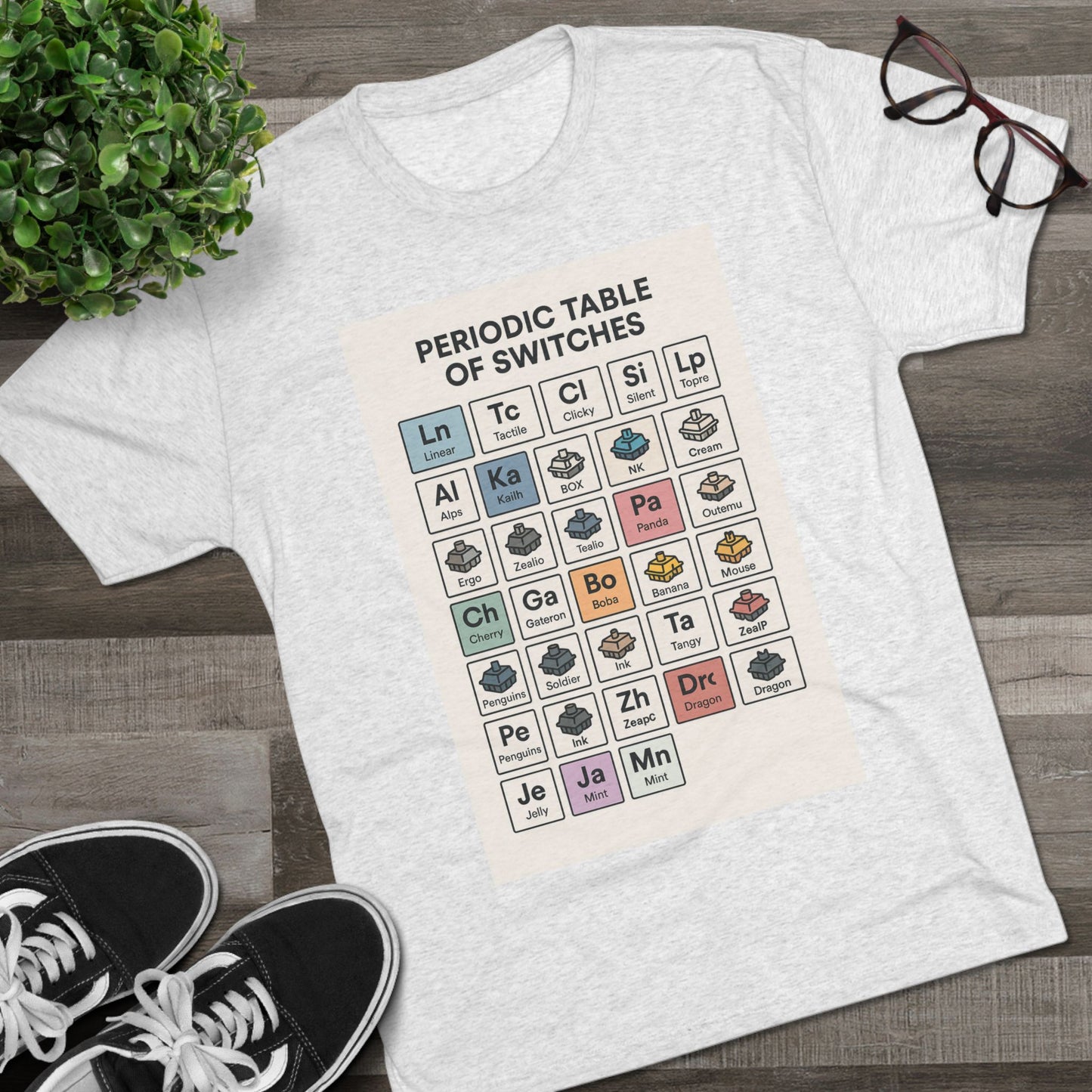 Flat lay of a heather gray T-shirt with a Periodic Table of Switches design, styled with sneakers and a plant — made for mechanical keyboard fans.