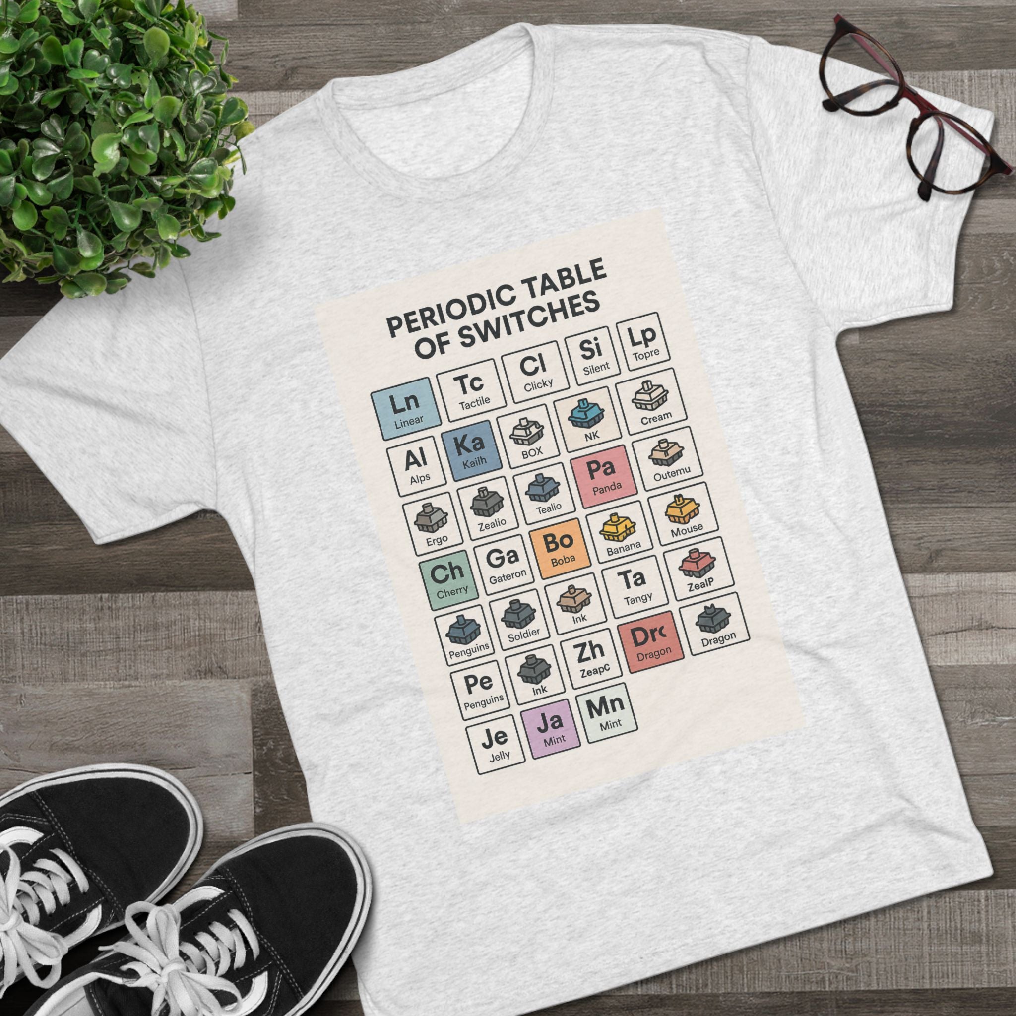 Flat lay of a heather gray T-shirt with a Periodic Table of Switches design, styled with sneakers and a plant — made for mechanical keyboard fans.